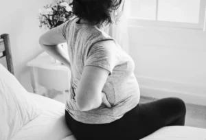 Pregnant woman sitting on bed holding lower back due to pain