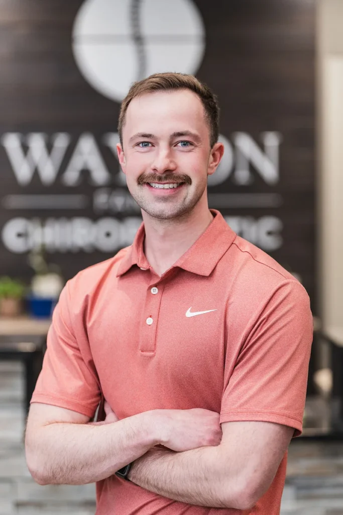 A smiling man with a mustache wearing a reddish-orange Nike polo shirt, with his arms crossed, standing in front of a blurred background with a 'Watson Chiropractic' logo.
