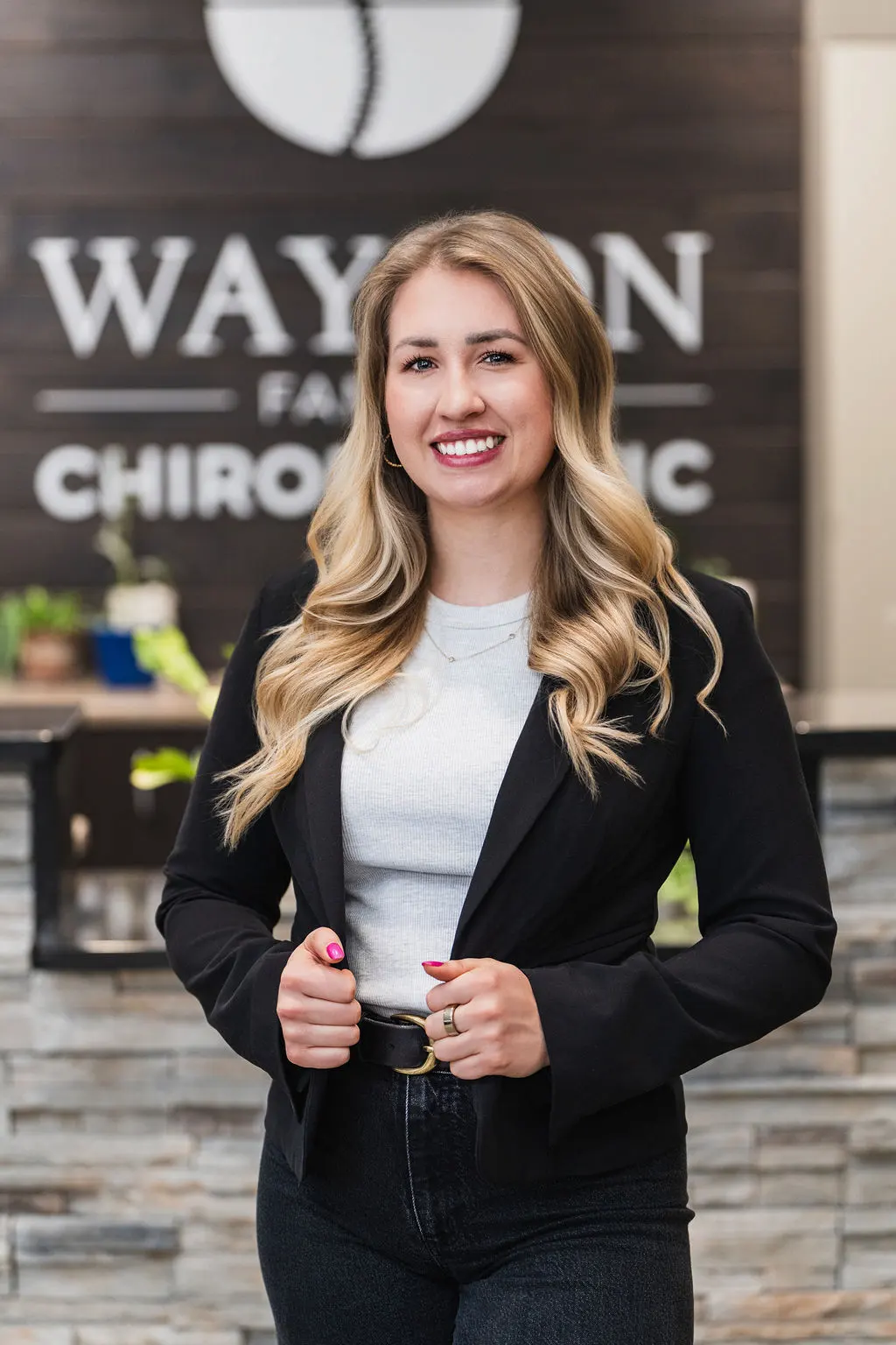 A smiling woman with blonde hair in a black blazer and grey top stands in front of a blurred chiropractic clinic sign.