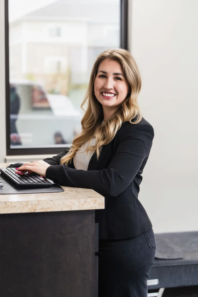 A smiling woman with blonde hair in a black blazer stands at a reception desk with her hands on a keyboard.