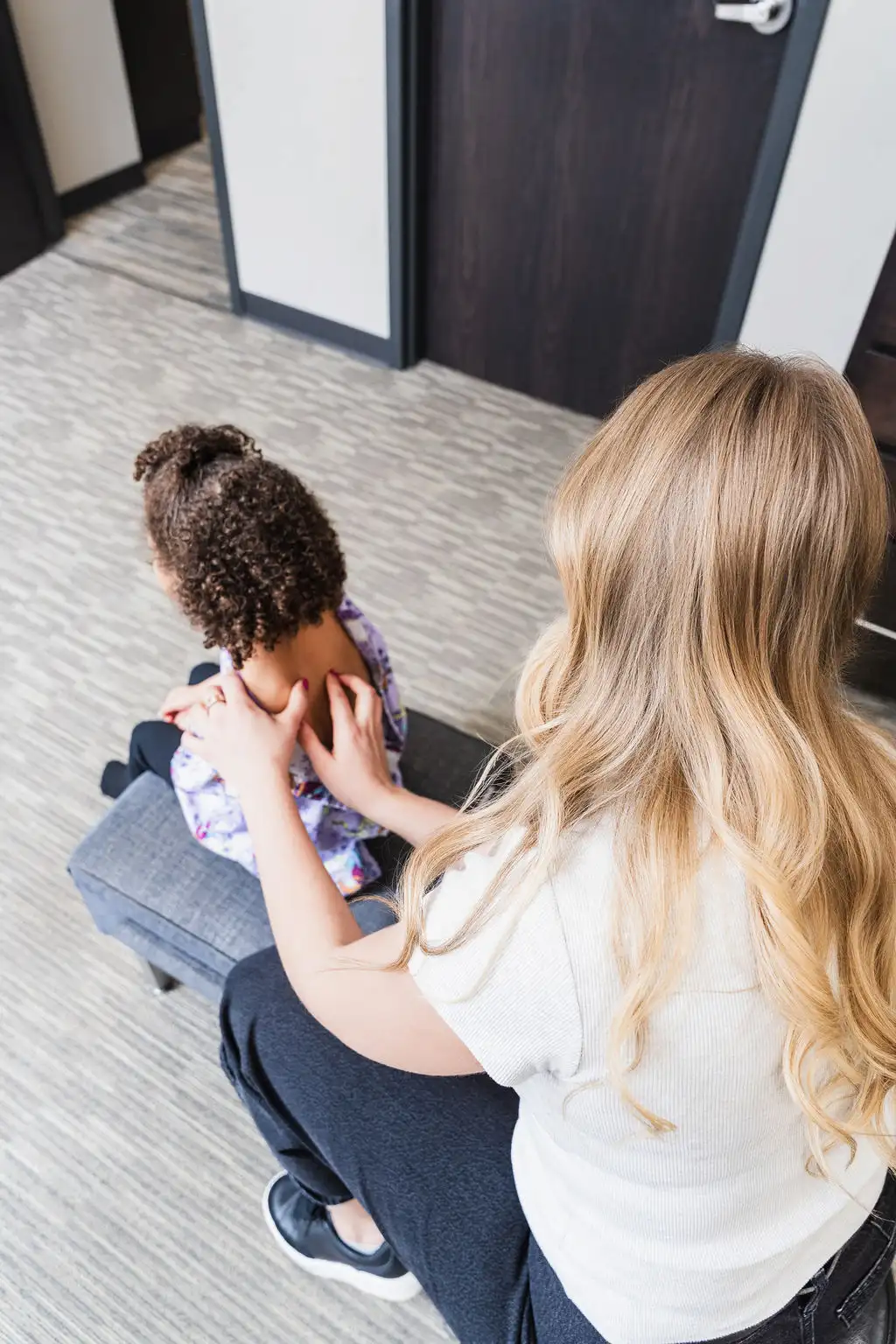 Overhead view of an adult with blonde hair massaging the upper back of a child with dark curly hair, who is sitting on a gray bench.
