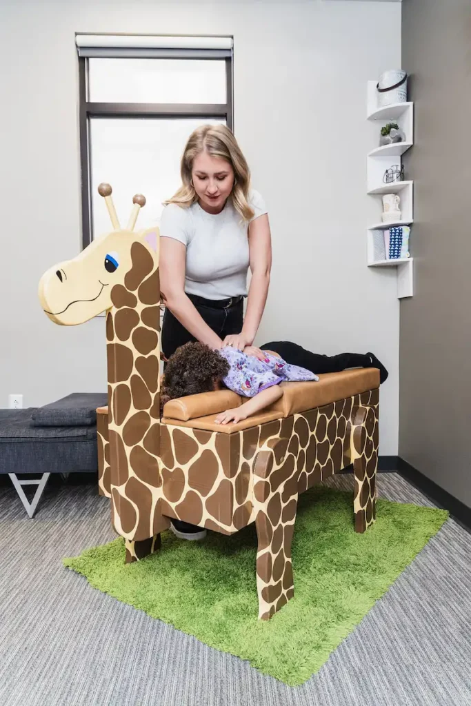 A woman adjusts a child lying on a giraffe-shaped chiropractic table in a child-friendly room.