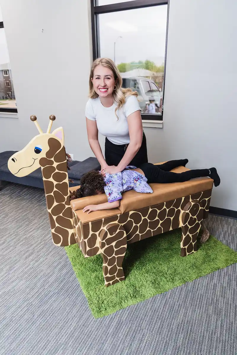 A smiling woman adjusts a child lying face down on a giraffe-shaped examination table in a bright room.