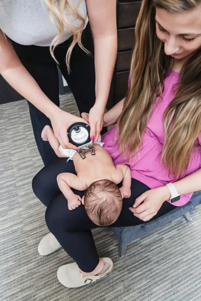 Two women with a baby. One woman holds the baby on her lap while another woman uses a medical device on the baby's back.