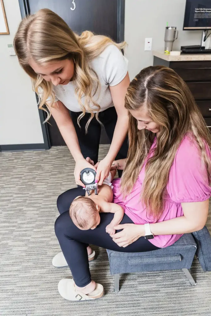 A chiropractor uses an instrument to examine a baby's back while the baby is held by a woman.