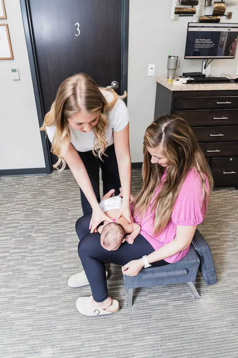 A chiropractor gently adjusts a newborn baby lying on its mother's lap in a clinic setting.