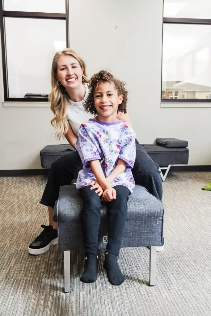 A smiling adult woman stands behind a young girl who is seated on a bench. Both are looking at the camera.