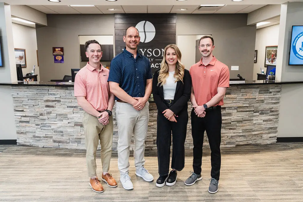 Four smiling staff members, three men and one woman, stand in front of a stone reception desk with the 'Grayson Family Chiropractic' logo behind them.