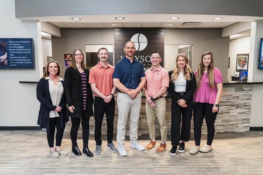 Seven people, four women and three men, standing in a row in a modern office reception area with a sign that says 'Wyson Academy' behind them.